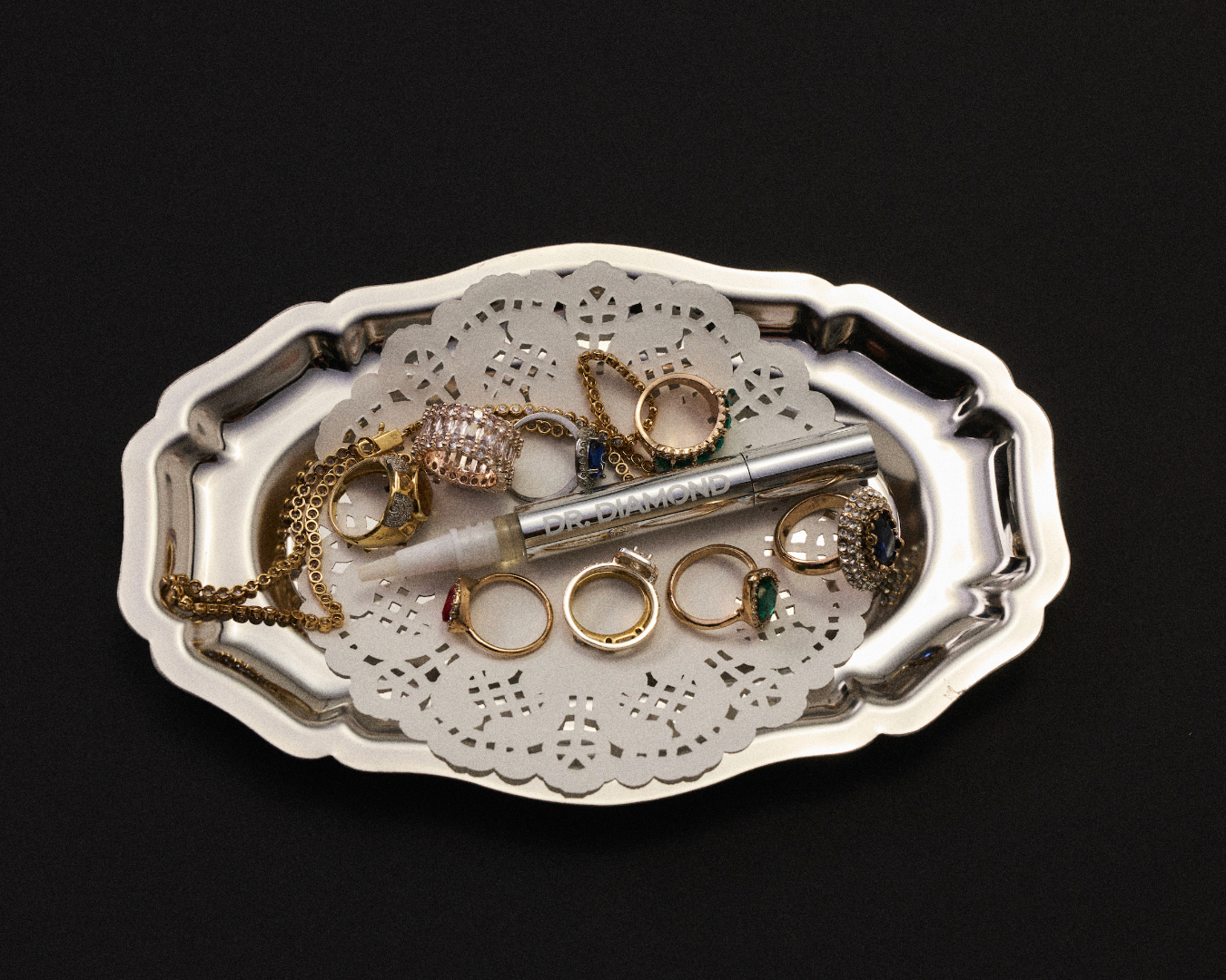 A silver-toned metal jewelry organizer tray with a scalloped edge, holding a white lace doily, several gold rings, and a silver pen, set against a solid black background.