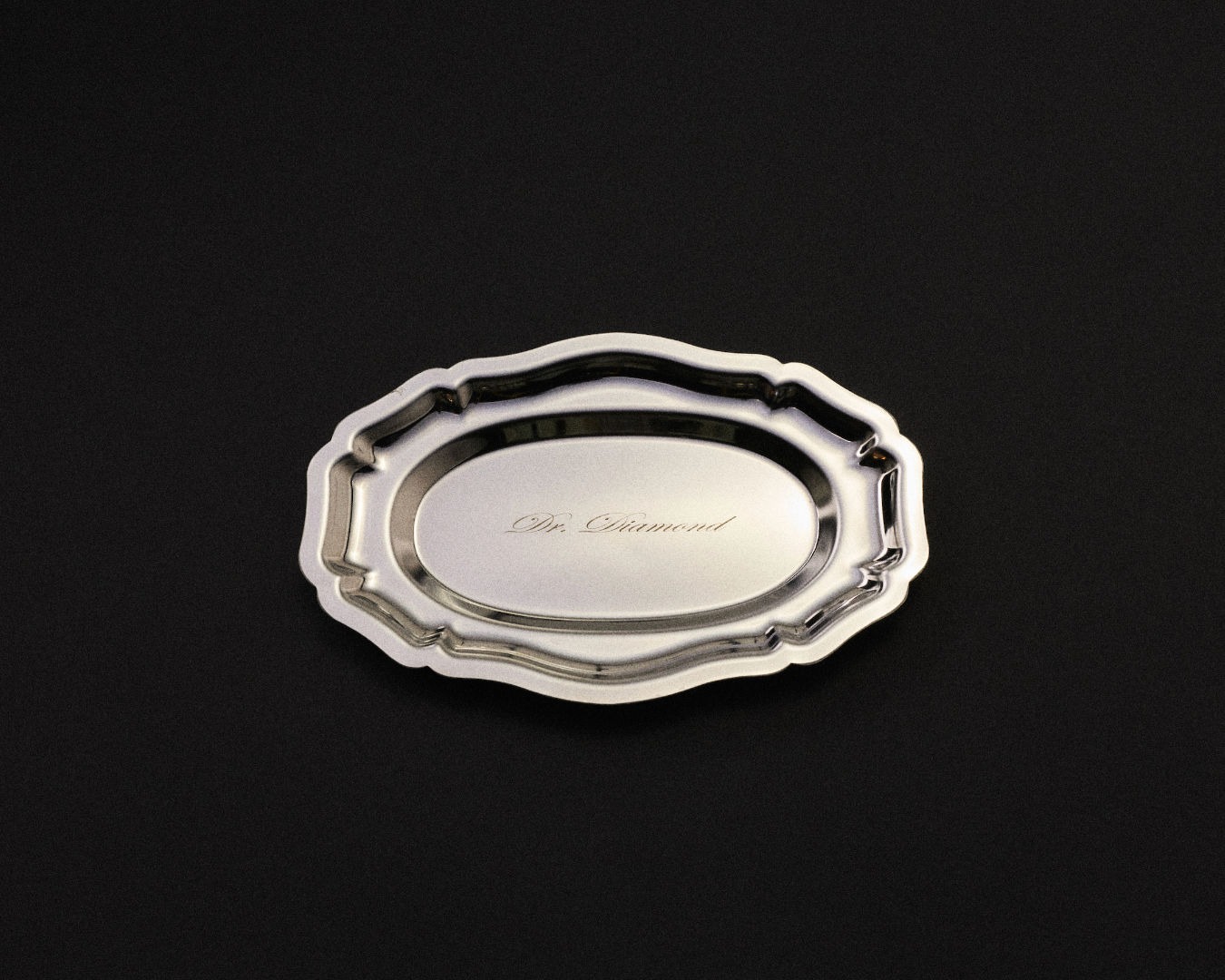 A polished silver oval tray with decorative scalloped edges and the engraved name 'Dr. Diamond' in the center, shown on a solid black background.