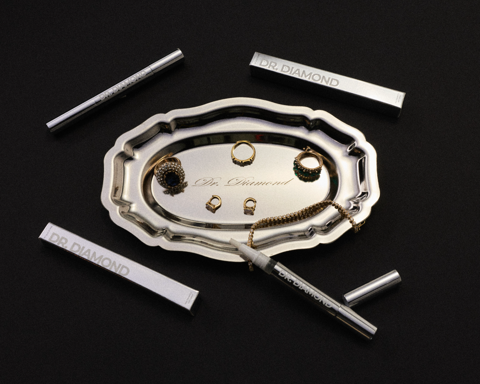 A silver-toned jewelry organizer tray with scalloped edges, surrounded by four slim silver cylindrical cases engraved with 'DR DIAMOND' on a black background. The central tray holds several pieces of gold and silver jewelry, including rings, earrings, and a chain.